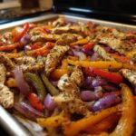 Close-up of seasoned chicken strips mixed with colorful bell peppers and red onions fresh from the oven for One-Pan Chicken Fajitas with Peppers and Onions.