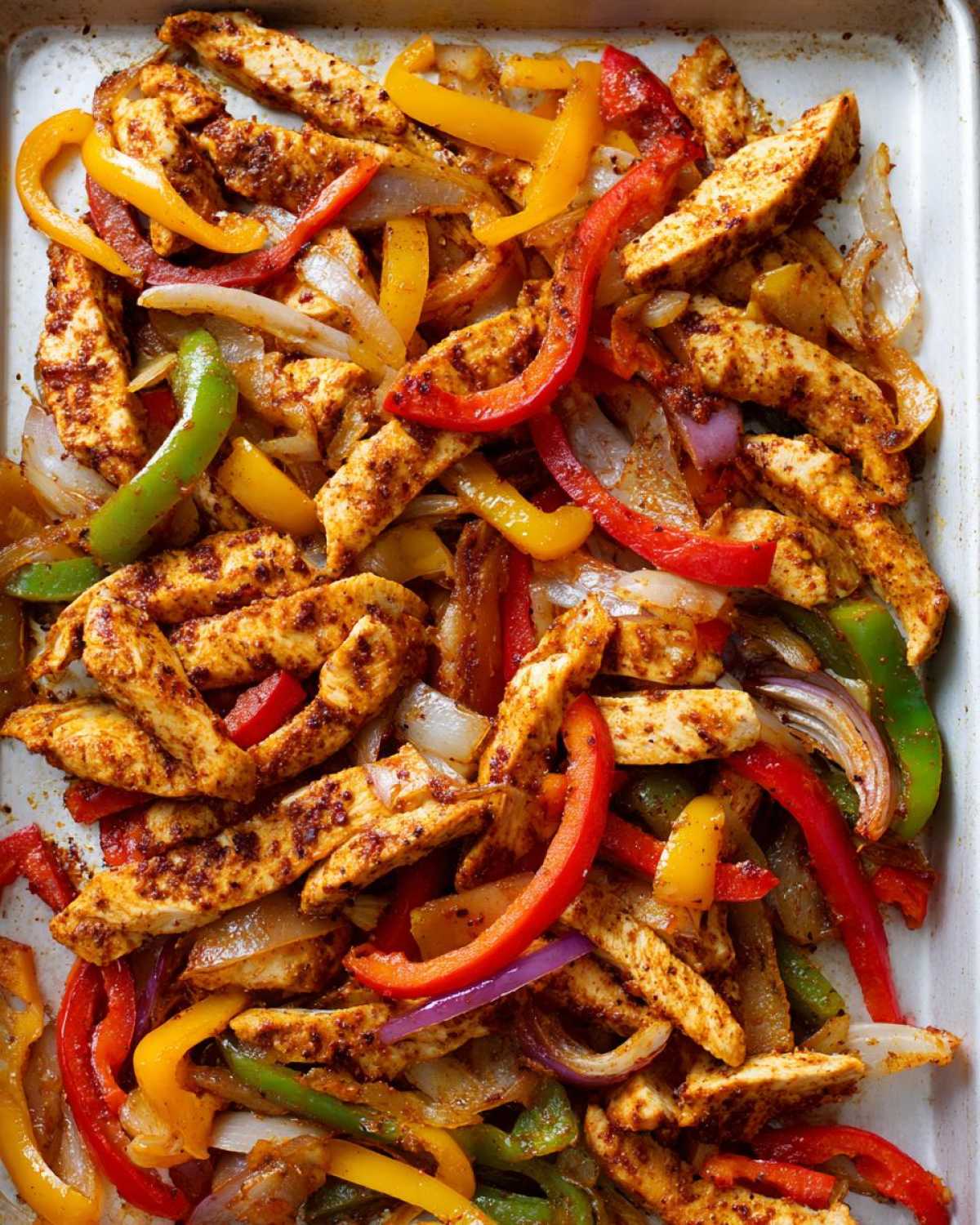 Close-up of seasoned chicken strips mixed with colorful bell peppers and onions cooked on a sheet pan for One-Pan Chicken Fajitas.