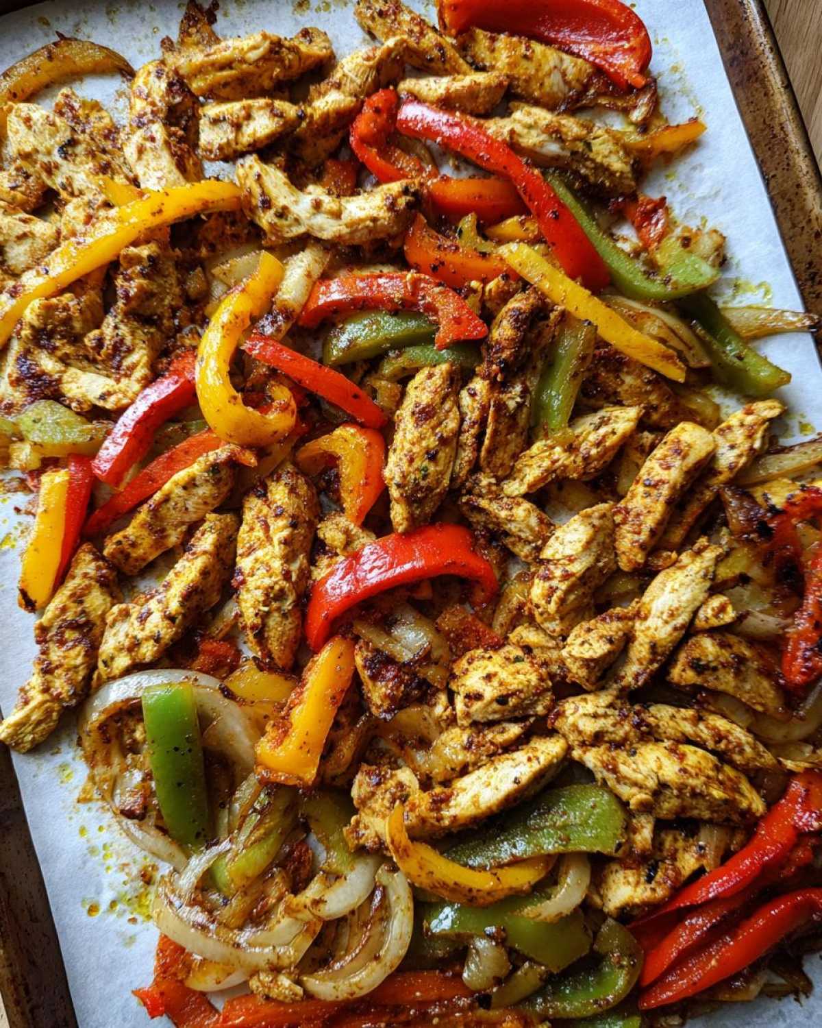 Close-up of seasoned chicken strips mixed with colorful bell peppers and onions cooked on a baking sheet for One-Pan Chicken Fajitas.