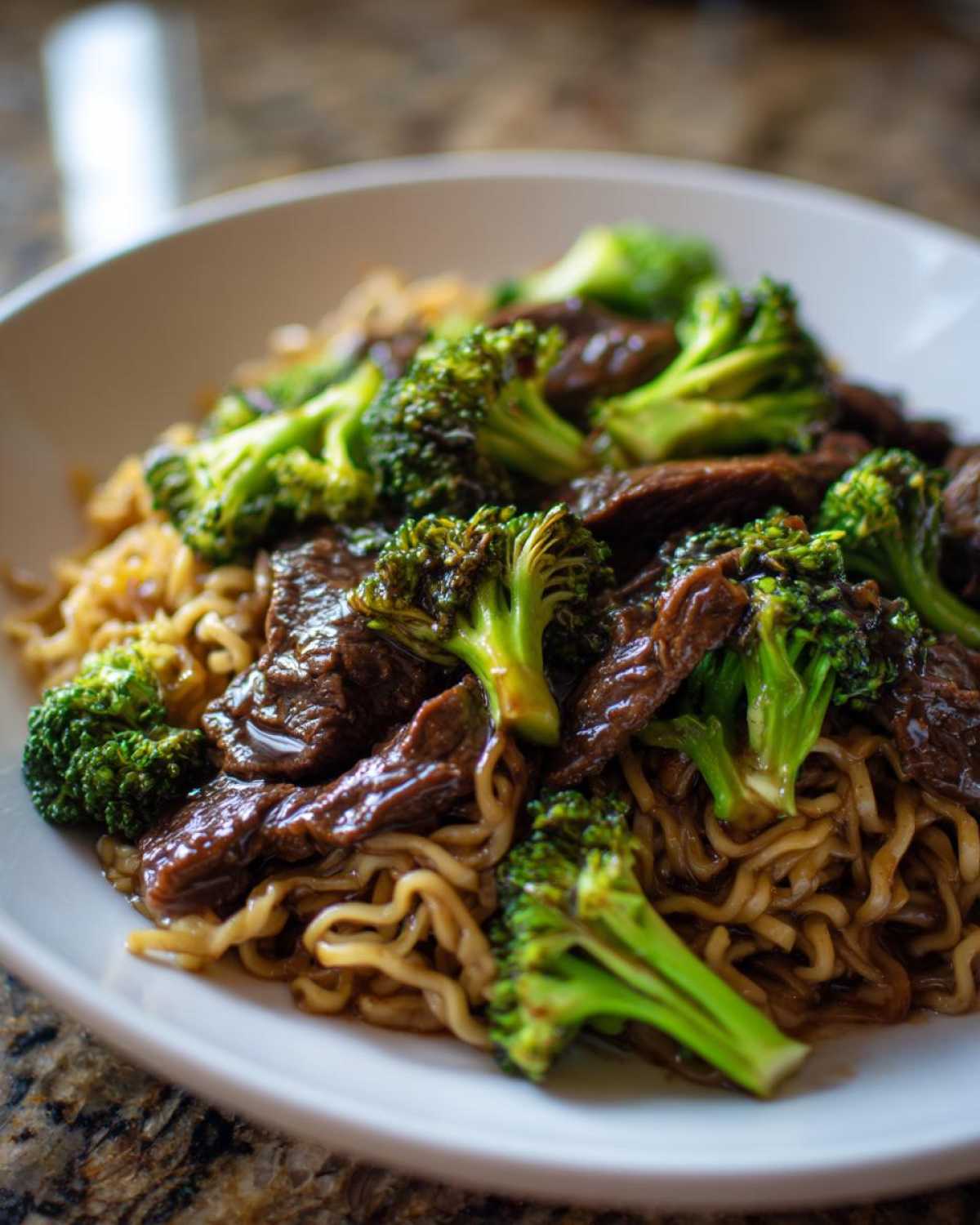 A close-up shot of a white bowl filled with One-Pot Beef and Broccoli Ramen Noodles, featuring glossy beef strips and bright green broccoli florets.