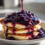 A stack of fluffy pancakes being generously topped with thick Homemade Blueberry Syrup and whole berries.