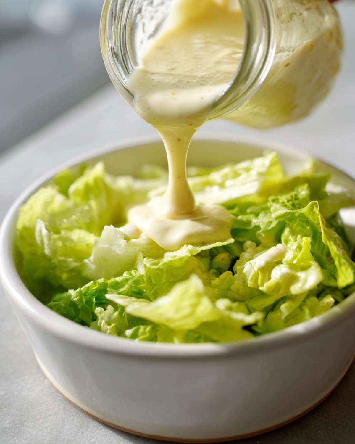 Creamy Copycat Olive Garden Salad Dressing being poured from a glass jar onto fresh, crisp romaine lettuce in a white bowl.