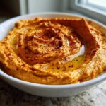 A close-up of creamy Roasted Red Pepper Hummus swirled in a white bowl, drizzled with olive oil and sprinkled with paprika.