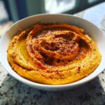 Close-up of creamy Roasted Red Pepper Hummus swirled in a white bowl, drizzled with olive oil and sprinkled with spices.