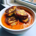 A white bowl filled with vibrant orange Roasted Tomato Basil Soup topped with golden brown Grilled Cheese Croutons.