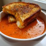 A white bowl filled with vibrant orange Roasted Tomato Basil Soup, topped with a grilled cheese sandwich cut into croutons.
