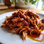 A close-up shot of shredded Slow Cooker Honey Garlic Pulled Pork coated in a thick, glossy sauce on a white plate.
