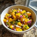 Close-up of a white bowl filled with bright yellow chunks of mango, red onion, and green herbs for Spicy Mango Salsa.