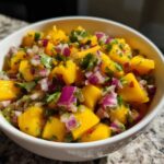 A close-up of a white bowl filled with bright yellow chunks of mango, red onion, and cilantro for Spicy Mango Salsa.
