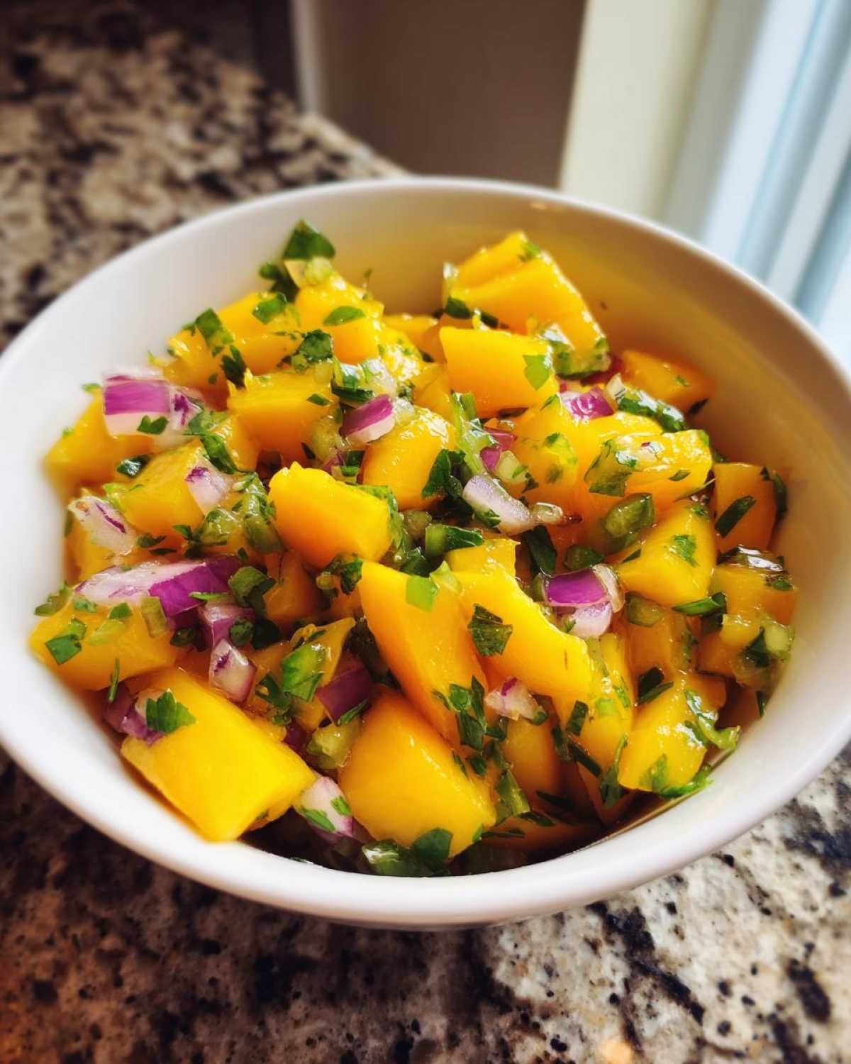 Close-up of a white bowl filled with bright yellow chunks of mango, red onion, and green cilantro for Spicy Mango Salsa.