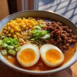 A bowl of Spicy Miso Ramen featuring soft-boiled eggs, ground meat, corn, and scallions in a rich broth.