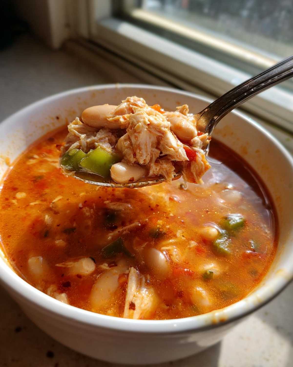 A spoonful of Spicy Turkey White Bean Chili showing shredded turkey, white beans, and green peppers lifted from a white bowl.