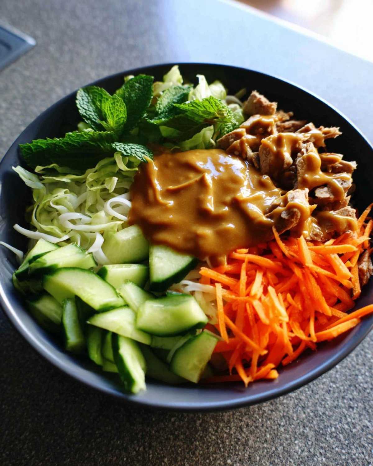 Close-up of a Spring Roll Bowl with Peanut Sauce, featuring shredded carrots, cucumbers, mint, and protein.