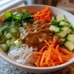 A close-up of a Spring Roll Bowl with Peanut Sauce, featuring rice noodles, shredded carrots, diced cucumber, and fresh mint.