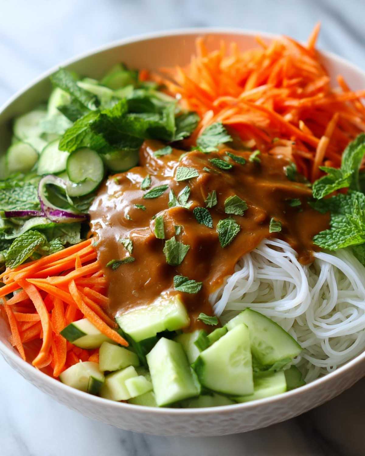 Close-up of a Spring Roll Bowl with Peanut Sauce poured over rice noodles, cucumbers, carrots, and mint.