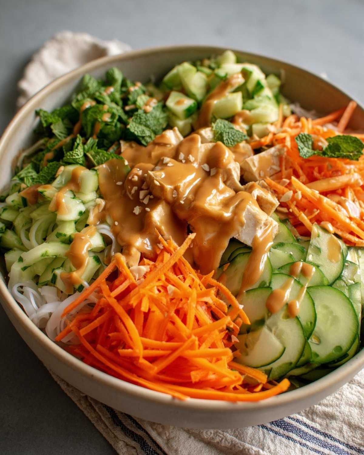 A vibrant Spring Roll Bowl with Peanut Sauce featuring rice noodles, shredded carrots, cucumbers, tofu, and fresh mint.