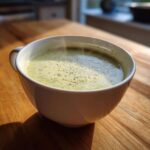 A steaming white mug filled with bright green Creamy Potato Leek Soup, garnished with herbs, sitting on a wooden table.