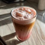 A glass of layered Strawberry Cream Cold Brew with pink foam topping, sitting on a sunlit marble counter.