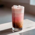 A tall glass showing distinct layers of dark cold brew coffee, pink strawberry cream, and thick white foam of the Strawberry Cream Cold Brew.