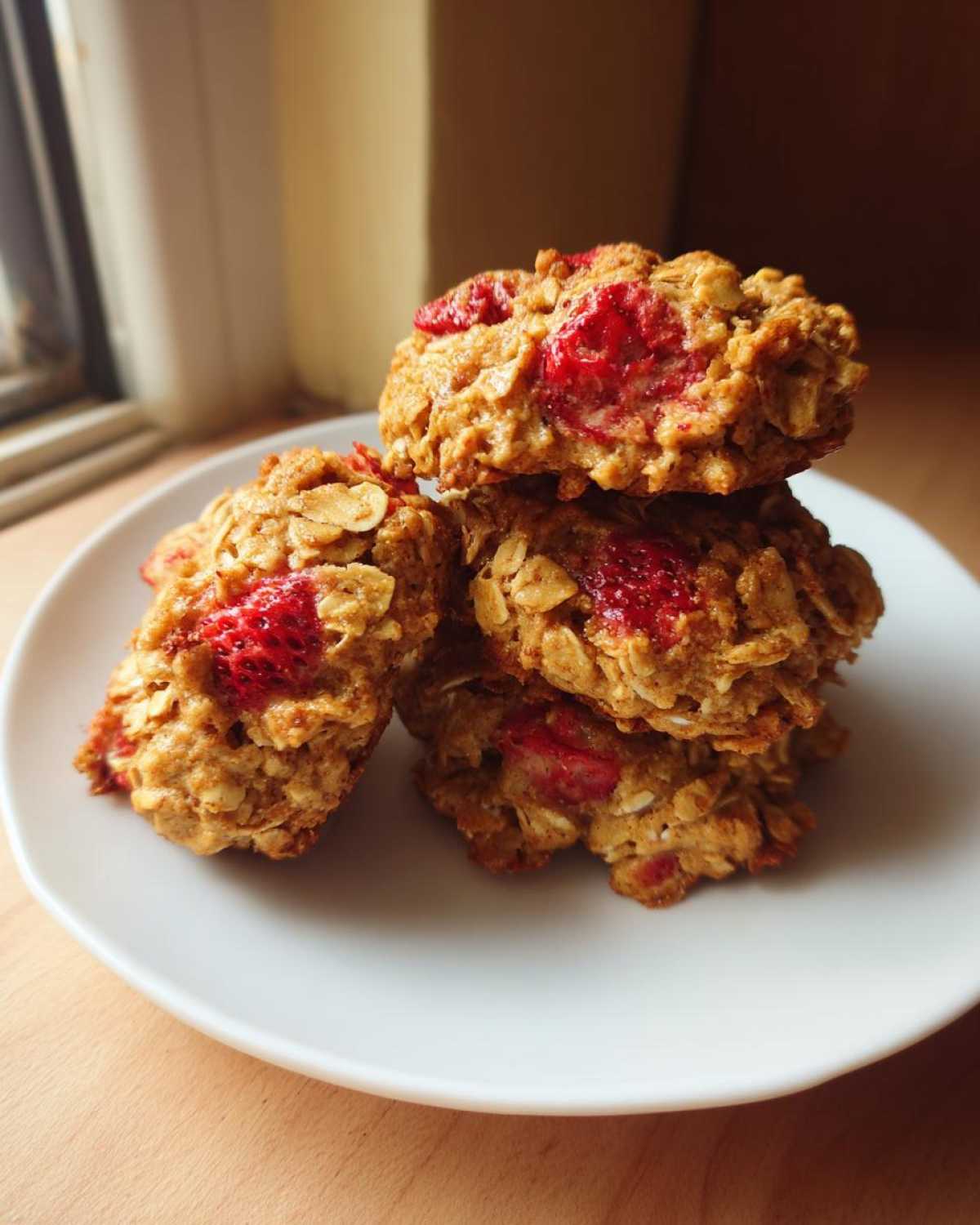 A stack of four golden Strawberry Oatmeal Breakfast Cookies studded with bright red fruit pieces on a white plate.