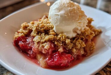 A close-up of a warm serving of Strawberry Rhubarb Crisp topped with a scoop of melting vanilla ice cream.