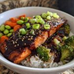 Close-up of a Teriyaki Salmon Rice Bowls featuring glazed salmon, white rice, broccoli, edamame, and carrots, topped with green onions.