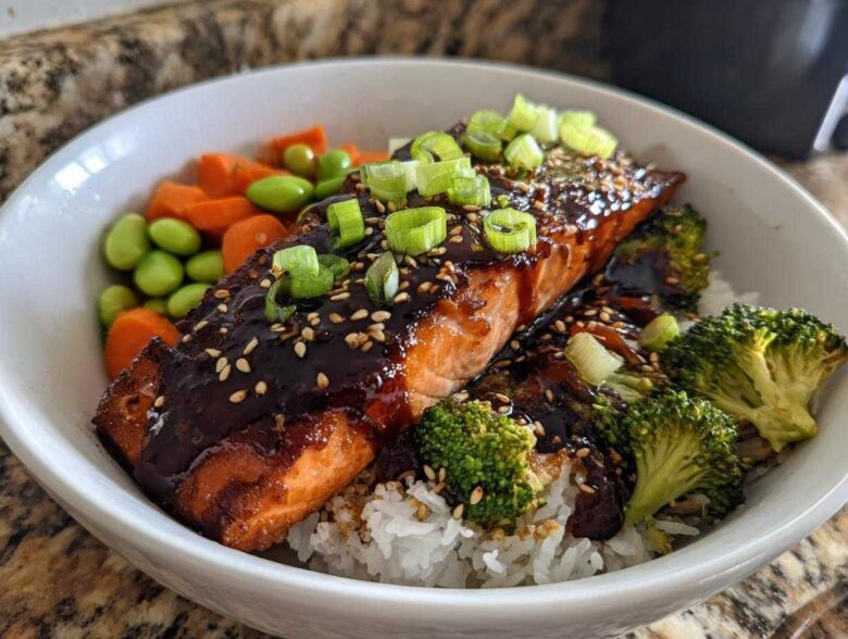 Close-up of a Teriyaki Salmon Rice Bowls featuring glazed salmon, white rice, broccoli, edamame, and carrots, topped with green onions.