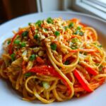A close-up of a bowl of creamy Thai Peanut Noodles with Crunchy Veggies, topped with chopped peanuts and scallions.
