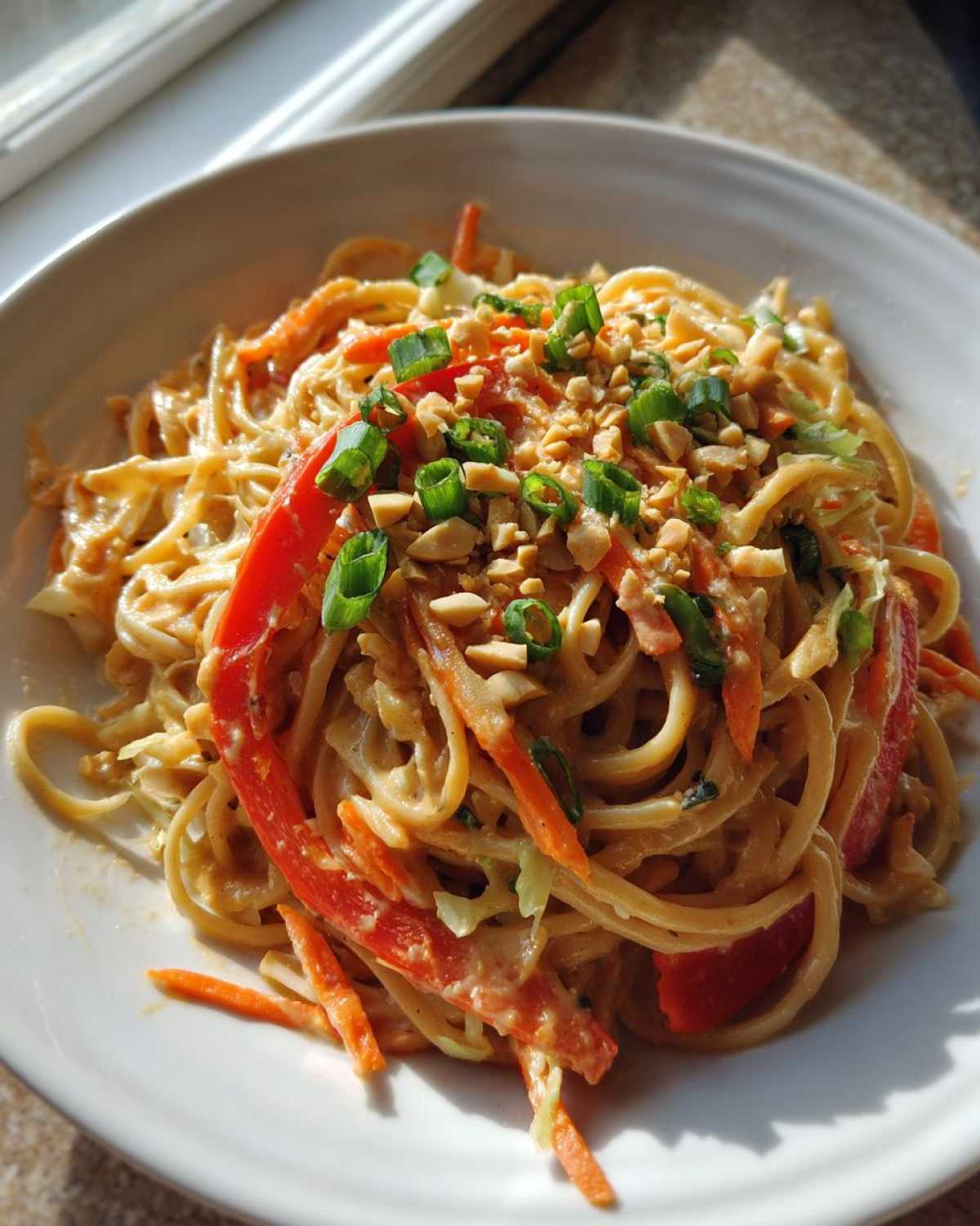 A white bowl filled with creamy Thai Peanut Noodles with Crunchy Veggies, topped with chopped peanuts and green onions.