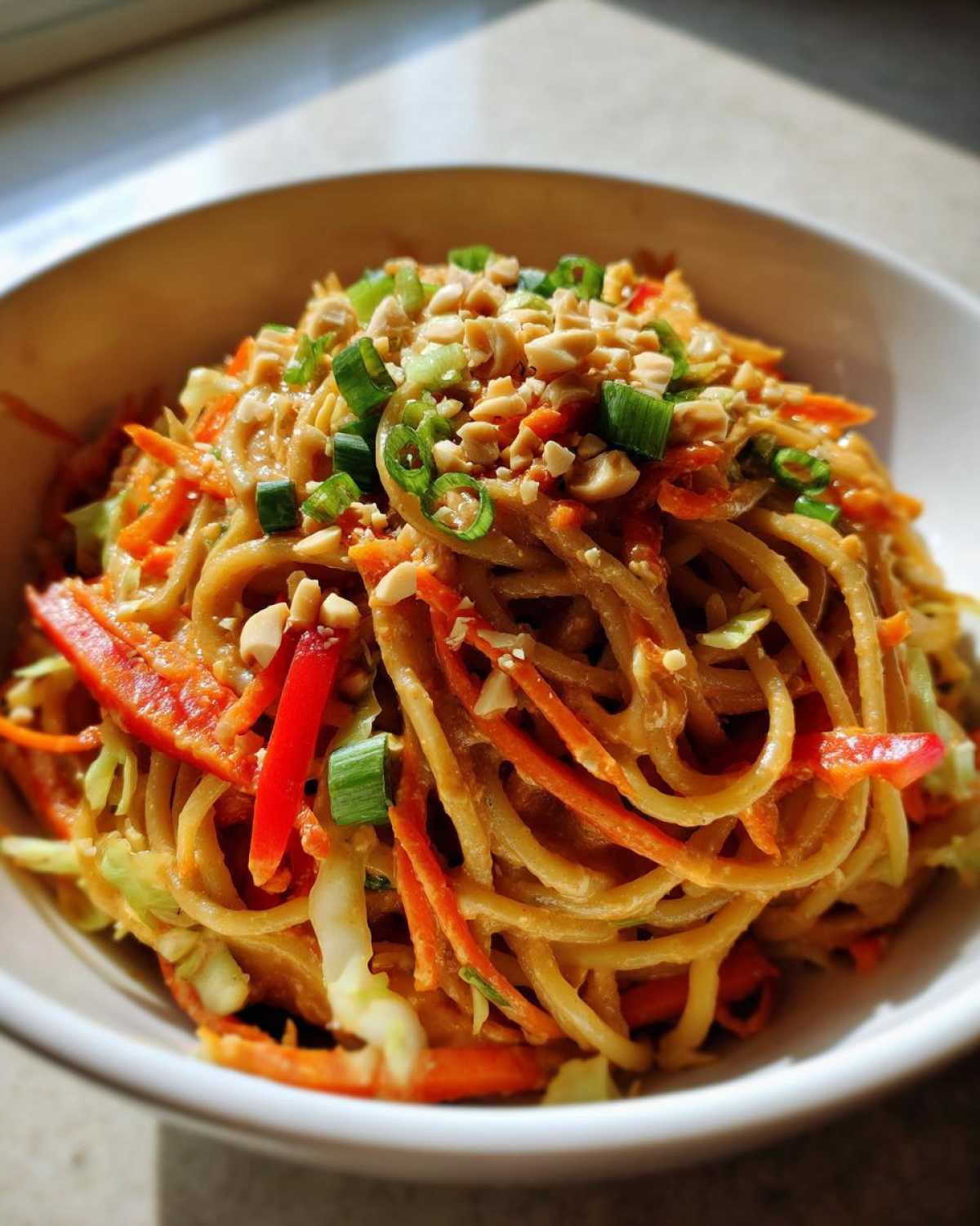 Close-up of a bowl of Thai Peanut Noodles with Crunchy Veggies, topped with chopped peanuts and green onions.