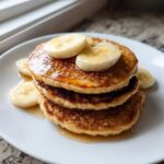 A stack of three fluffy Vegan Banana Oat Pancakes drizzled with syrup and topped with fresh banana slices.