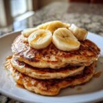 A stack of three fluffy Vegan Banana Oat Pancakes topped with sliced bananas and drizzled with syrup.