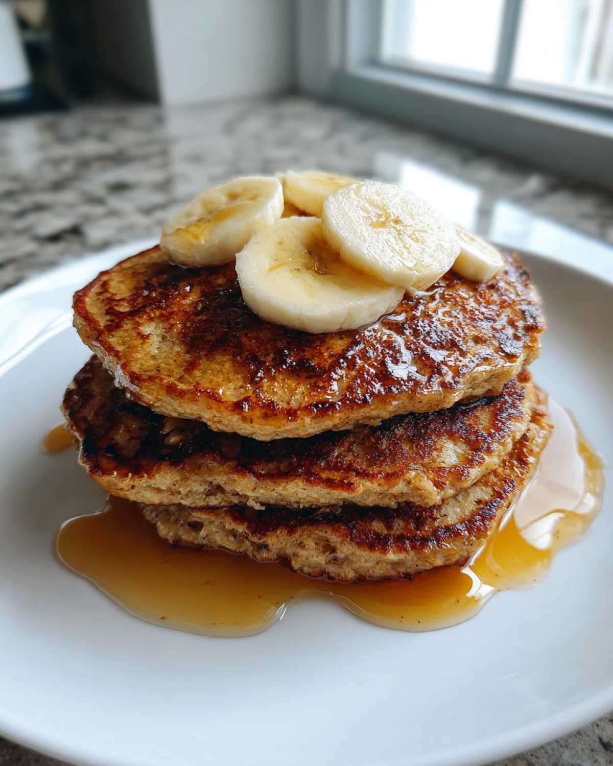 A stack of three fluffy Vegan Banana Oat Pancakes drizzled with syrup and topped with fresh banana slices.