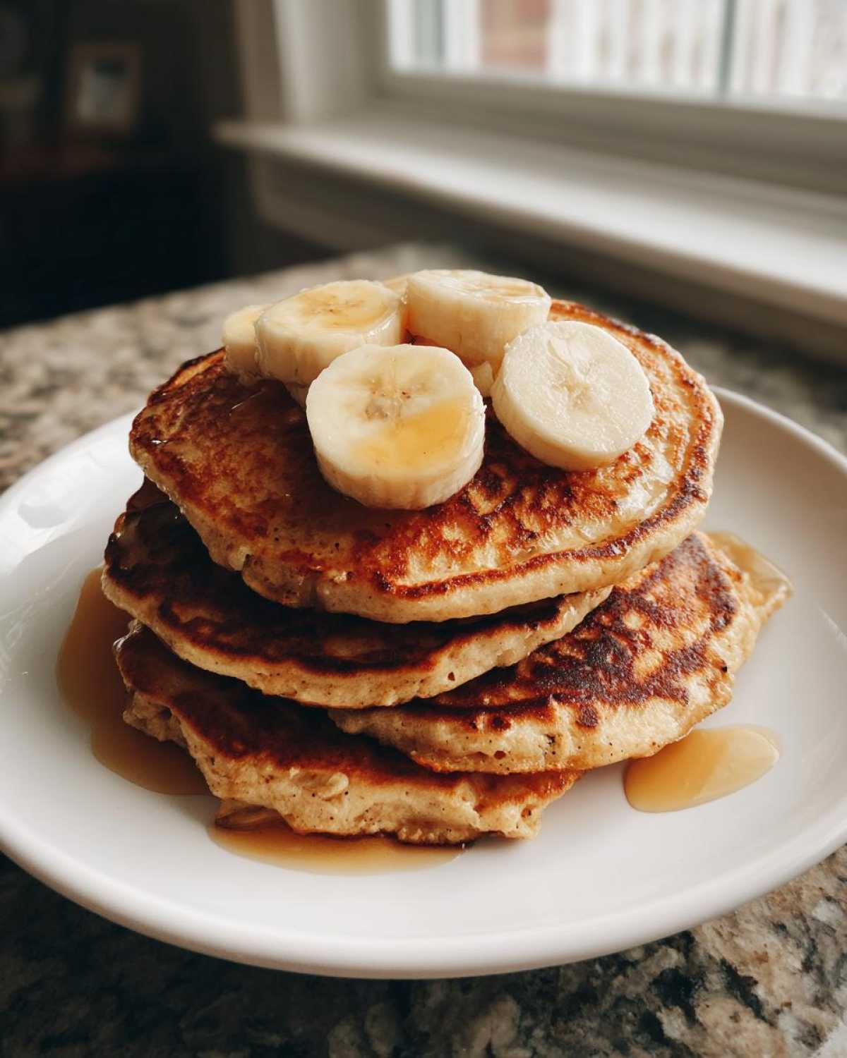 A stack of fluffy Vegan Banana Oat Pancakes topped with sliced bananas and drizzled with maple syrup.