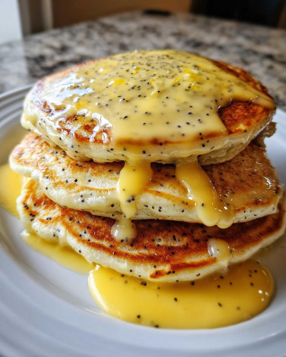 A stack of three fluffy Vegan Lemon Poppy Seed Pancakes drizzled with bright yellow lemon glaze.
