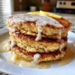 A tall stack of fluffy Vegan Lemon Poppy Seed Pancakes drizzled with glaze and topped with lemon slices.