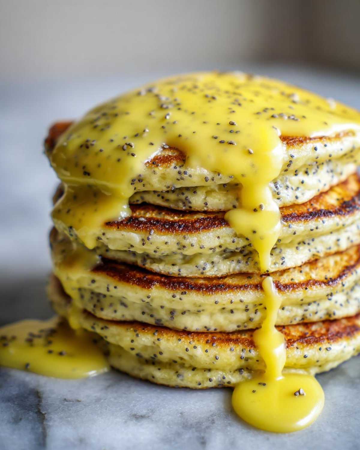 Close-up of a tall stack of fluffy Vegan Lemon Poppy Seed Pancakes topped with a thick, dripping lemon glaze and poppy seeds.