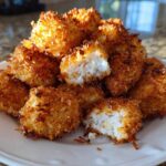 A pile of golden brown Air Fryer Crispy Coconut Chicken Bites on a white plate, one is broken open showing the white chicken inside.