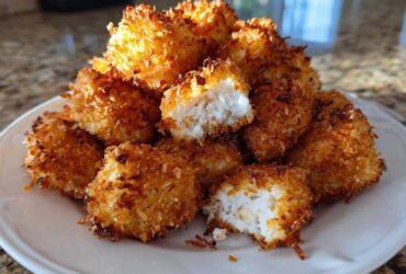 A pile of golden brown Air Fryer Crispy Coconut Chicken Bites on a white plate, one is broken open showing the white chicken inside.