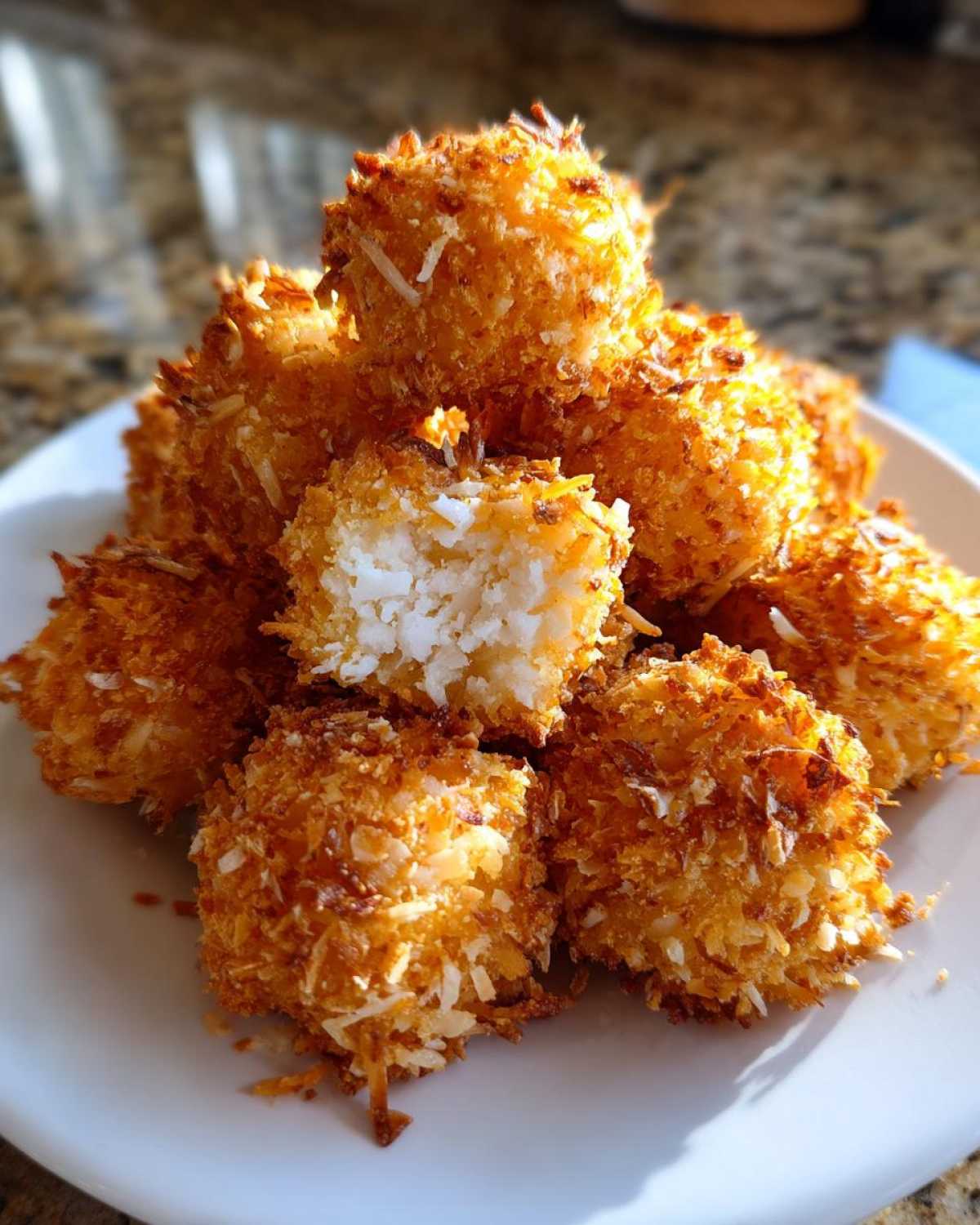 A pile of golden brown Air Fryer Crispy Coconut Chicken Bites on a white plate, one is broken open showing white chicken inside.