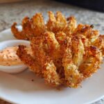Two golden brown Air Fryer Crispy Onion Petals served on a white plate next to a small bowl of spicy mayo dipping sauce.