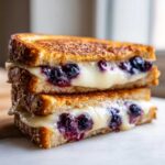 Close-up of two stacked halves of a golden-brown Blueberry Grilled Cheese sandwich with melted white cheese and bursting blueberries.