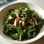 A vibrant bowl of Copycat Kale Crunch Salad tossed with sliced almonds and seeds, glistening under natural light.