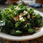 A vibrant close-up of Copycat Kale Crunch Salad tossed with dressing and topped with toasted sliced almonds on a white plate.