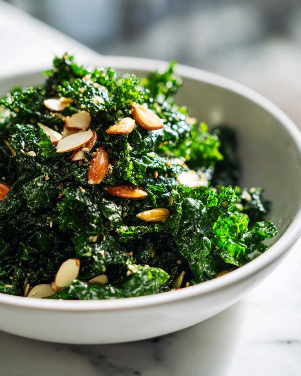 Close-up of vibrant green kale tossed in dressing and topped with slivered almonds in a white bowl, featuring the Copycat Kale Crunch Salad.