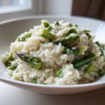 A close-up of a serving of Easy Creamy Asparagus Risotto, featuring creamy rice mixed with bright green asparagus pieces and peas, presented in a white bowl.