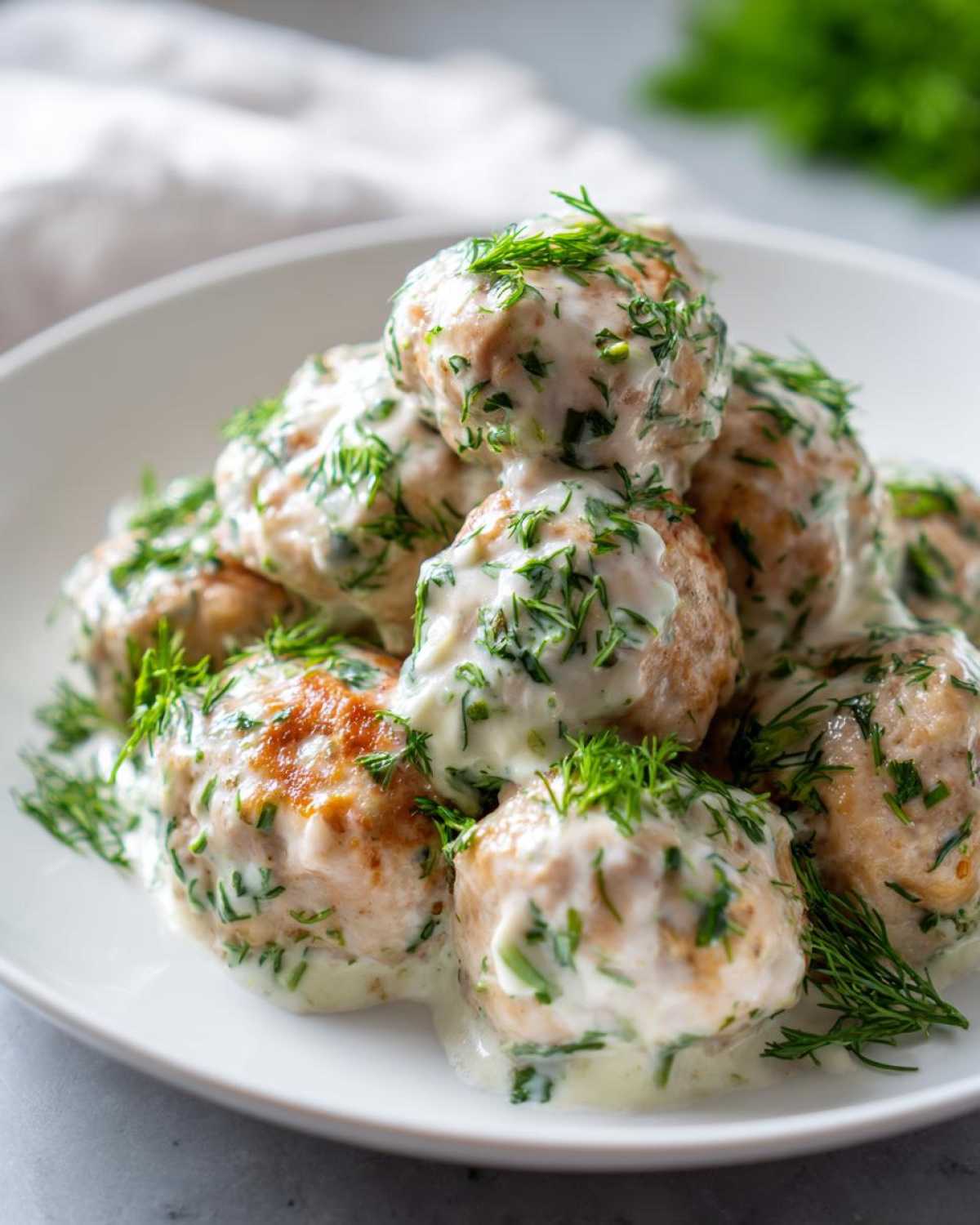 A mound of Easy Creamy Greek Chicken Meatballs covered in a white dill sauce, served on a white plate.