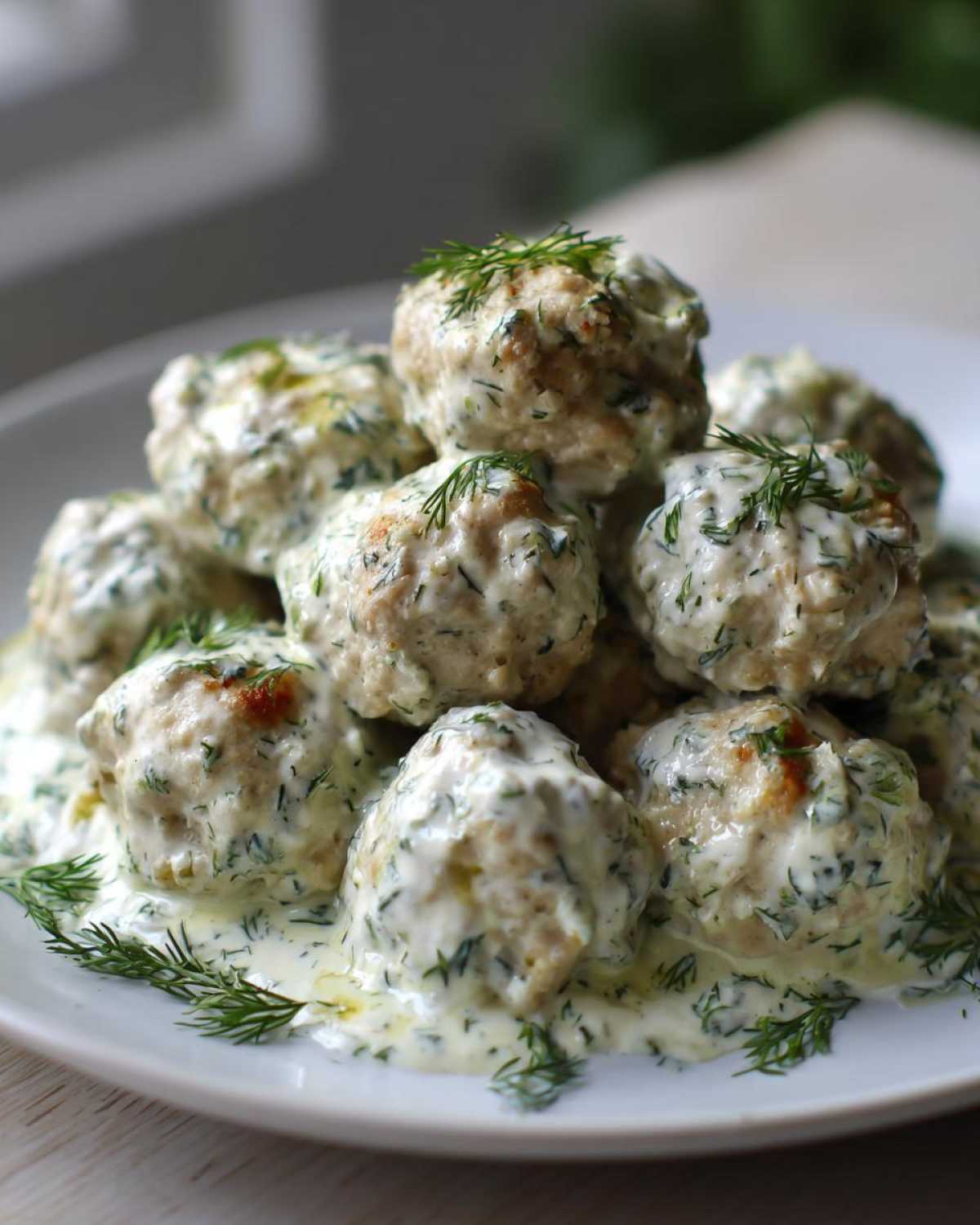 A mound of Easy Creamy Greek Chicken Meatballs covered in a rich, dill-flecked cream sauce on a white plate.