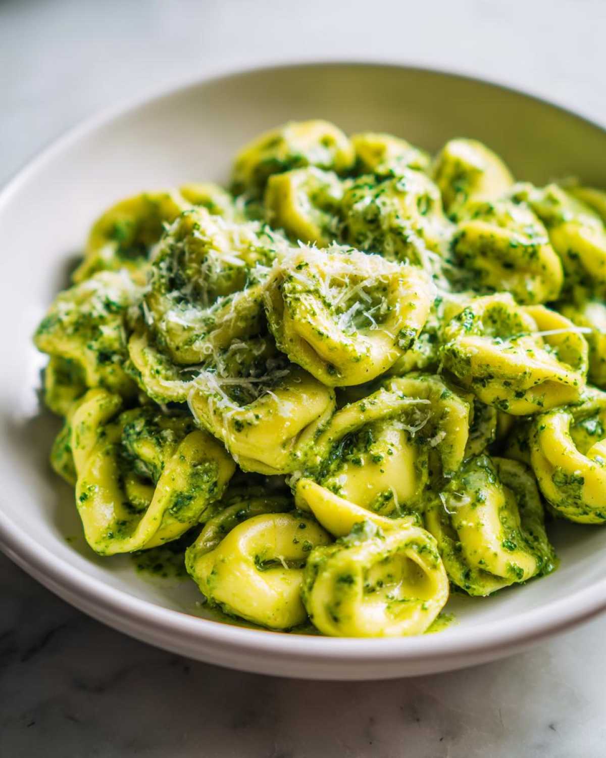 A close-up of a white bowl filled with Easy One-Pot Creamy Pesto Tortellini topped with grated Parmesan cheese.