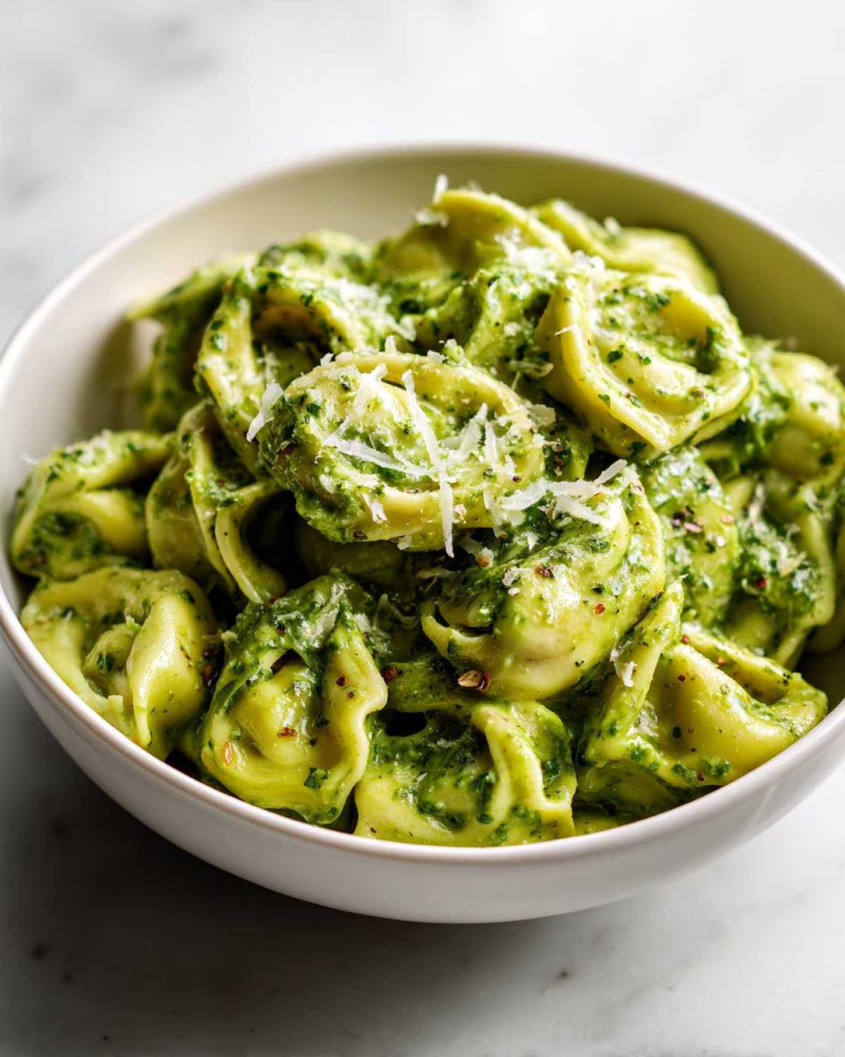 A white bowl filled with Easy One-Pot Creamy Pesto Tortellini topped with grated Parmesan cheese and red pepper flakes.